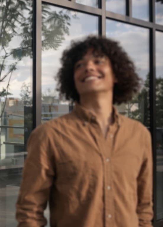 Blurry smiling young man with dark curly hair wearing a brown button-down shirt outdoors.